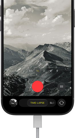 iPhone 18 Pro mostrato di fronte, sul display un paesaggio montuoso, con fotocamera in modalità Time-Lapse e cavo di alimentazione collegato alla porta di ricarica